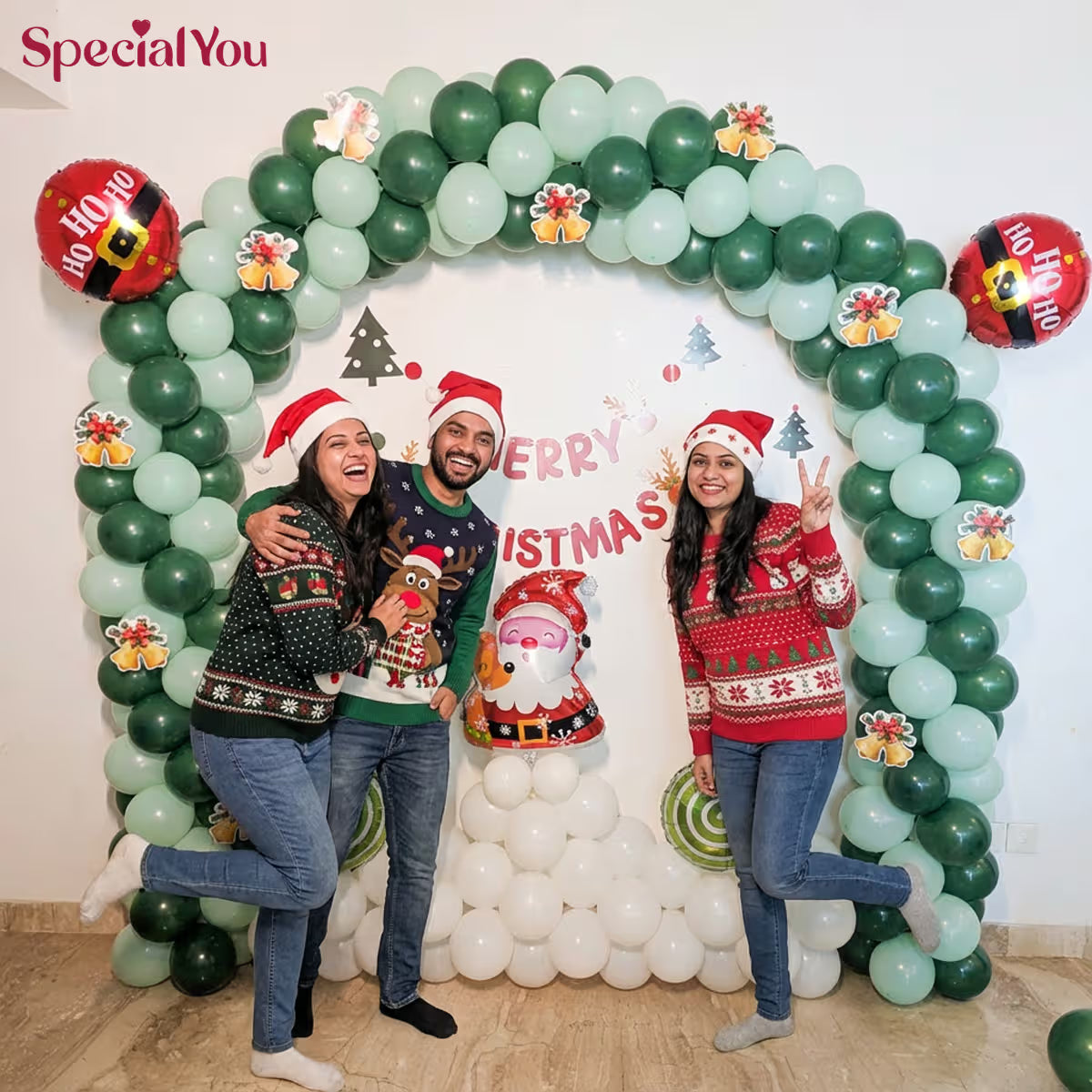 Three people in Christmas sweaters posing through a festive balloon arch with 'Merry Christmas' text.